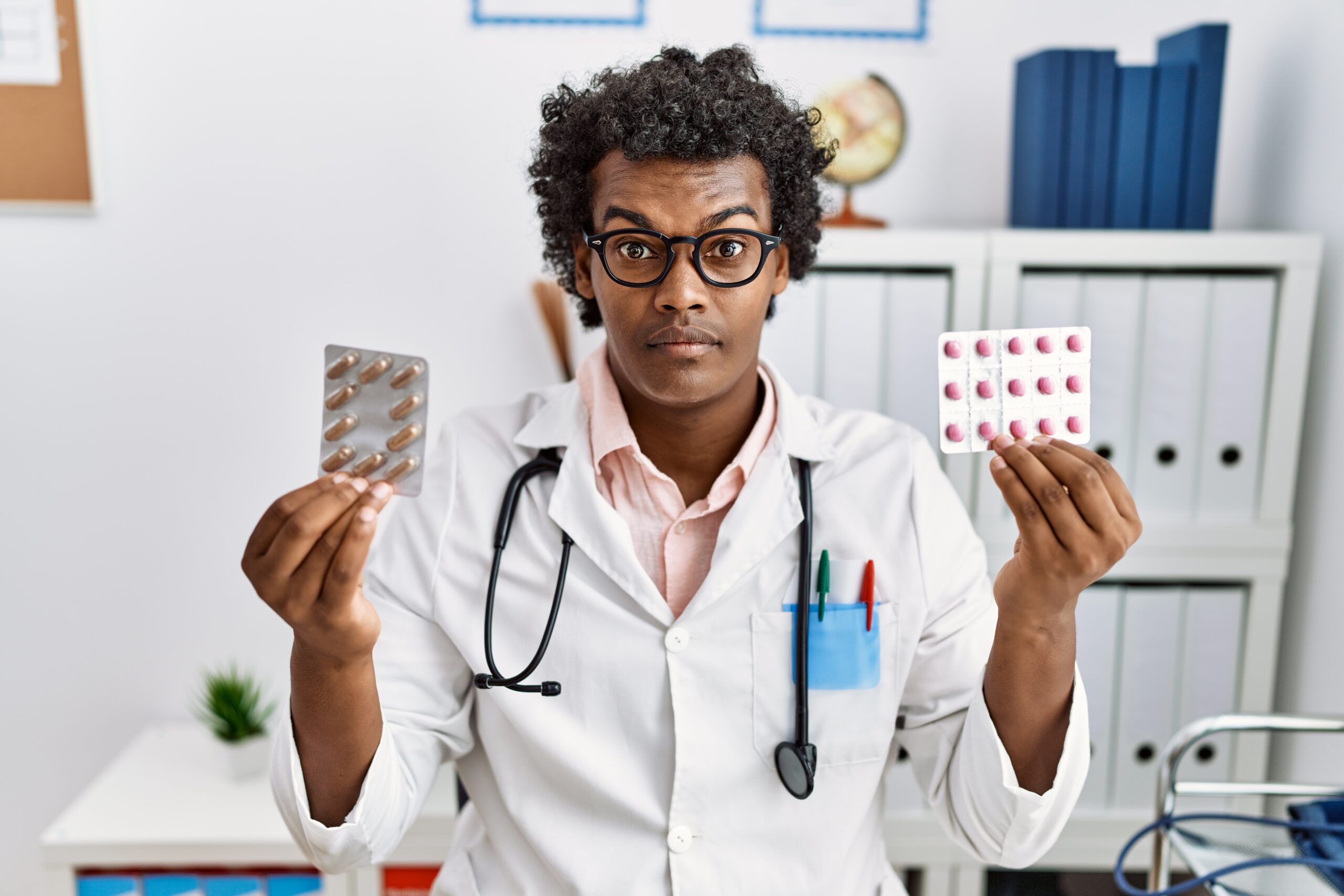 African man wearing doctor uniform holding prescription pills in shock face, looking skeptical and sarcastic, surprised with open mouth